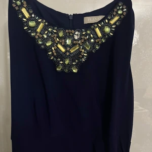 Mörkblå långklänning med stenar från Planet - Snygg mörkblå långklänning med dragkedja i ryggen. Halsringningen är dekorerad med stora glittrande stenar i grönt och gult. Klänningen har drapering på sidan.