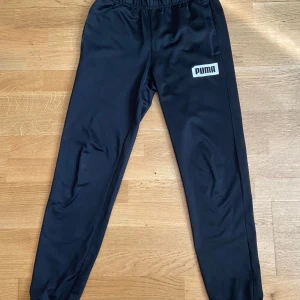 Svarta träningsbyxor från Puma - Svarta mjukisbyxor från Puma med vit logga på vänster lår. Byxorna har vit snörning och är i glansigt polyestermaterial. Okej skick-har pytterlitet hål på ett knä-syns knappt i livet och omöjligt i kamera. 