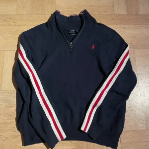 Half  zip tröja Polo Ralph Lauren - Marinblå stickad tröja från Polo Ralph Lauren med half zip och klassisk röd logga på bröstet. Snygga vita och röda ränder längs ärmarna ger en unik stil. Tröjan är väldigt ovanlig då den inte längre säljs.