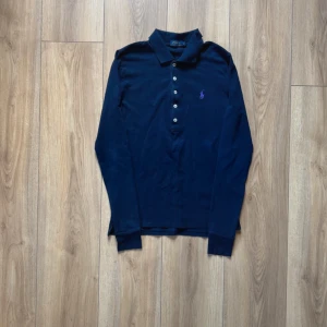 Marinblå långärmad piké från Polo Ralph Lauren - Snygg marinblå långärmad pikétröja från Polo Ralph Lauren med klassisk krage och knappar framtill. Tröjan har ett lila broderat logotyp på bröstet och är gjord i mjuk bomull. Perfekt för dig som gillar stilrena och tidlösa plagg.