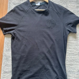 Svart Gymshark t-shir - Svart t-shirt från Gymshark med broderad logga på bröstet. Klassisk rund halsringning och korta ärmar. Tillverkad i mjuk bomull, perfekt för gymmet eller chill. Enkel och stilren design som funkar till det mesta. Liten defekt bakom tröjan, man kan sy ihop det enkelt därav priset 