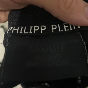 Svart Philipp Plein henley med rosa logga - Svart henley-skjorta från Philipp Plein med korta ärmar och knappar vid halsen. På bröstet sitter en rosa PP-logga i ett sexkantigt emblem. Skjortan har en clean och modern look, perfekt för dig som gillar streetwear och exklusiva märken.