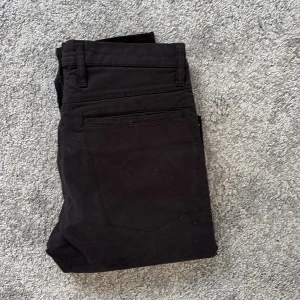 Svarta byxor från Nudie Jeans, 30/34 - Svarta byxor från Nudie Jeans i klassisk rak modell. Byxorna har diskreta bakfickor och bälteshällor, tillverkade i bomull med en stilren look. Perfekta för dig som gillar enkel och clean stil.