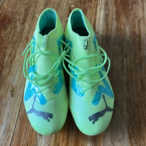 Puma limegröna fotbollsskor - Snygga limegröna fotbollsskor från Puma med blå detaljer och svarta Puma-loggor. Skorna har snörning och mönstrad ovandel i syntetmaterial samt dobbar i gult och blått för bra grepp på planen. Perfekta för dig som vill sticka ut på matchen!