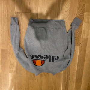 Grå sweatshirt från Ellesse XS - Snygg grå sweatshirt från Ellesse i storlek XS. Tröjan har klassisk logga framtill i svart, orange och rött. Mjuk insida och ribbade muddar vid ärmslut och midja. Perfekt för en chill och sportig stil.