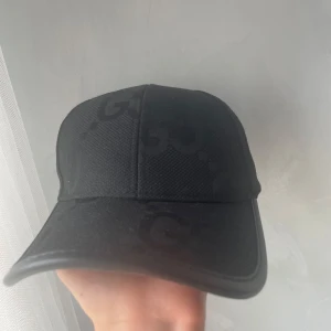 Svart keps från Gucci med GG-mönster - Snygg svart keps från Gucci med diskret GG-logga i samma färg över hela kepsen. Klassisk böjd skärm och justerbar baksida. Tillverkad i ett tåligt tygmaterial som ger en exklusiv känsla. Perfekt accessoar för att lyfta din outfit.