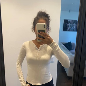 Vit ribbad långärmad topp med knappar - Crèmefärgad långärmad topp med ribbad struktur och knappdetaljer framtill. Modellen är figurnära med rund halsringning och vågiga kanter. 