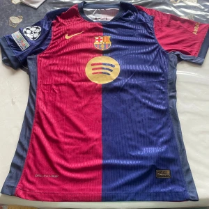 FC Barcelona Raphinha 11 matchtröja - Säljer en officiell FC Barcelona matchtröja från Nike med Raphinha 11-tryck. Tröjan har klassisk halv röd och halv blå design, guldiga detaljer och klubbmärke på bröstet. Tillverkad i lätt och ventilerande material med korta ärmar och tryckta sponsorer.