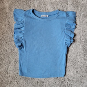 Blå ribbad topp med volangärmar Zara - Supersöt blå ribbad topp från Zara med volangdetaljer på axlarna. Toppen är ärmlös med breda volanger som ger en extra touch. Perfekt för dig som gillar en enkel men ändå unik stil. Liten i storleken, passar som en storlek S.