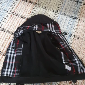 Svart hoodiezip från Burberry - Svart hoodiezip från Burberry med dragkedja, klassiskt rutigt foder i huvan och broderad logga på bröstet. Jackan har snörning i huvan och ribbade muddar. Perfekt för dig som vill ha en stilren och lyxig streetwear-look.