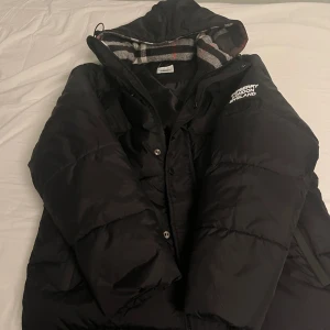 Svart pufferjacka från Burberry - Säljer en snygg svart pufferjacka från Burberry med huva och knappar. Jackan har ett stilrent Burberry London England-märke på armen och ett klassiskt rutigt foder. Perfekt för kyliga dagar!
