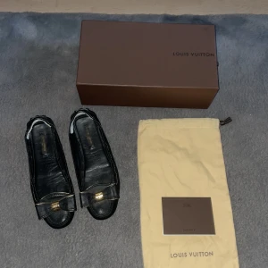 Svarta ballerinaskor Louis Vuitton - Stilrena svarta ballerinaskor från Louis Vuitton i skinn med dekorativ rosett framtill och guldfärgad LV-detalj. Skorna har rund tå, kontrastsömmar och platt sula med gummidetaljer undertill. Perfekta för dig som gillar lyxiga detaljer och klassisk design.