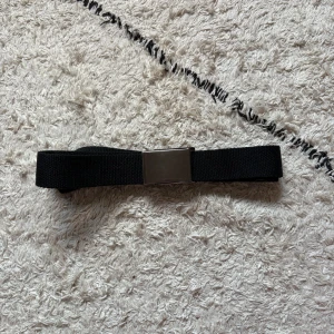 Svart tygbälte med metallspänne - Enkel och stilren svart tygbälte med ett rektangulärt metallspänne. Bältet är gjort i slitstarkt material och passar perfekt till jeans eller chinos för en clean look. Spännet är i silverfärg och ger en modern touch.