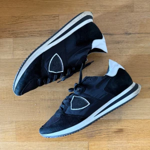 Philippe Model TRPX - Svarta sneakers från Philippe Model med vita detaljer på hälen och sula. Skorna har en blandning av mocka och textil, klassisk snörning och den ikoniska sköld-loggan på sidan. Snygg, sportig look med rund tå och platt sula.