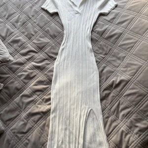 Vit ribbad långklänning från NA-KD - Supersnygg vit ribbad långklänning från NA-KD med krage, v-ringning och korta ärmar. Klänningen har en figurnära passform och en slits nertill för extra edge. Perfekt för dig som gillar stilrena och trendiga plagg.