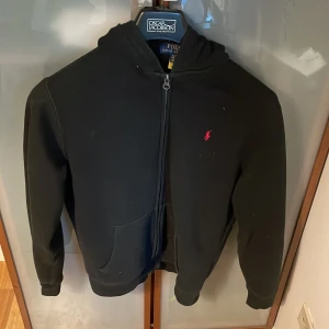 Svart hoodie från Polo Ralph Lauren - Svart hoodie från Polo Ralph Lauren i storlek XL i barn/ xs i vuxen med dragkedja och huva. Klassisk röd broderad logga på bröstet och känguruficka framtill. Mjuk och skön bomullsblandning, perfekt för chill dagar eller när du vill ha en clean look. 