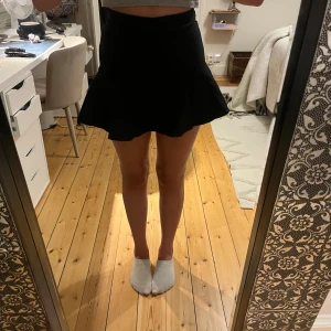 Svart volangkjol med knytband - Supersöt svart kortkjol med volangkant och knytband i midjan. Kjolen har en lätt utsvängd form som ger en snygg siluett och passar perfekt till crop tops eller stickade tröjor. Enkel att styla och ett måste i garderoben!