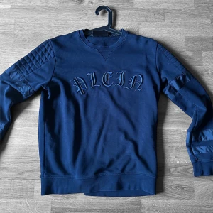 Blå sweatshirt från Philipp Plein - Säljer en marinblå sweatshirt från Philipp Plein med broderad logga framtill och coola quiltade detaljer på ärmarna. Tröjan har rund halsringning och är tillverkad i mjuk bomull. Perfekt för dig som gillar streetwear med lyxig känsla.