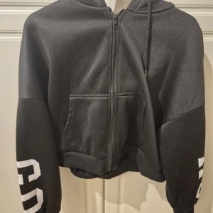 Svart hoodie ICA New York croppad - Svart croppad hoodie med vit text 'ICA New York EST 2015' på ryggen och tryck på ärmarna. Tröjan har huva med snörning, dragkedja framtill och en stor magficka. Perfekt för en chill och sportig look.