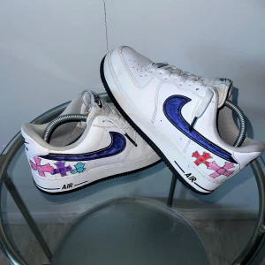 Nike Air Force 1 custom sneakers - Unika Nike Air Force 1 sneakers i vitt skinn med handmålade motiv i rosa, lila, rött och blått på hälen. Swoosh-loggan är markerad i mörkblått och sulan är svart. Klassisk låg modell med snörning och perforerad tå för extra stil.