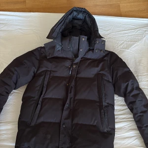 Svart dunjacka från Canada Goose S - Säljer en svart dunjacka från Canada Goose i storlek S. Jackan har huva, dragkedja och tryckknappar framtill, samt två stora fickor med dragkedja. Klassisk Canada Goose-logga på ärmen och quiltad design för extra värme. Perfekt för kalla dagar.