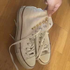 Vita Converse All Star med nitar - Klassiska vita Converse All Star high tops med coola guldfärgade nitar på sidan. Skorna har snörning, rund tå och är i canvas med gummisula. Perfekta för dig som vill ha en unik twist på dina sneakers.