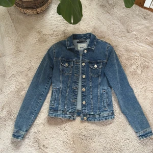 Blå jeansjacka från ONLY, strl 34 - Snygg klassisk blå jeansjacka från ONLY i storlek 34. Jackan har silverfärgade knappar, två bröstfickor med lock och två sidofickor. Har andvänts en gång 
