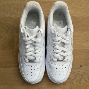 Nike Air Force 1 helvita sneakers - Klassiska Nike Air Force 1 sneakers i helvitt skinn med perforerad tå, rund tå och platt sula. Skorna har snörning och en metallbricka med 'AF1' på snörena. Perfekt clean look och ikonisk siluett som alltid är trendig.