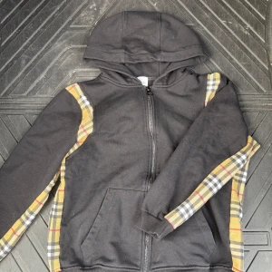 Svart Burberry hoodie med rutiga detaljer - Svart hoodie från Burberry med klassiska rutiga paneler i beige, rött och svart längs ärmar och sidor. Tröjan har huva, dragkedja framtill och känguruficka. Perfekt för dig som vill ha en stilren streetlook med lyxig touch.