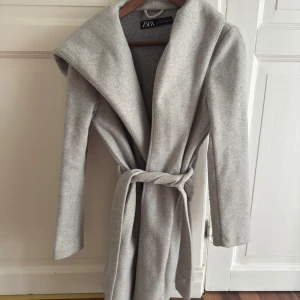 Grå kappa med huva från Zara - Snygg grå kappa från Zara med huva och bälte i midjan. Kappan har lång ärm och en clean, minimalistisk look som passar perfekt till lager-på-lager. Materialet är mjukt och känns lyxigt, och färgen är ljusgrå vilket gör den enkel att matcha.