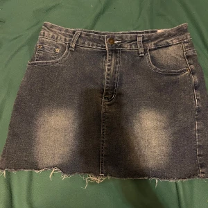 Svart jeanskjol med broderade fickor - Cool jeanskjol med slitna kanter och snygga broderade bakfickor med vita detaljer. Kjolen har klassisk femficksmodell, bälteshällor och en rå, fransig fåll för en edgy look. Perfekt för dig som gillar streetstyle och vill sticka ut. Endast använd 1 eller 2 gånger. Storlek 38 men kan nog  även passa storlek 36. 