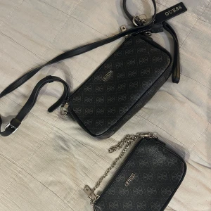Svart axelväska från Guess med kedja - Säljer en svart axelväska från Guess med logomönster och guldfärgade detaljer. Väskan har både justerbar axelrem i skinnimitation och en snygg kedjerem. Praktisk dragkedja upptill och flera fack inuti. Perfekt storlek för mobilen och det viktigaste. Väskan kommer med en liten guess necessär som man även kan ha i väskan 