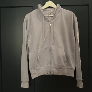 Ljuslila hoodie med dragkedja från H&M - En stilren ljuslila hoodie från H&M med dragkedja framtill, huva med snörning och stora fickor. Tröjan är långärmad och gjord i mjuk bomull, perfekt för chill dagar eller när du vill ha en avslappnad look.
