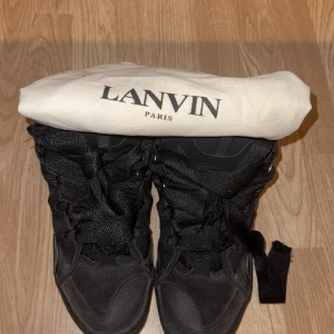 Svarta sneakers från Lanvin strl 45 - Säljer ett par svarta high-top sneakers från Lanvin i storlek 45.  Skorna har breda snören med fiskbensmönster, detaljer i både skinn och textil samt en rejäl platt sula. Snygg och chunky design, perfekt för dig som gillar statement-sneakers. ( använd ett par gånger)