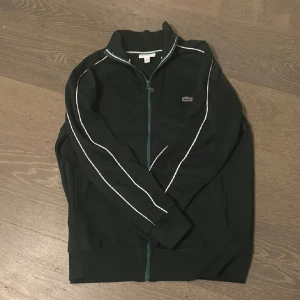 Mörkgrön track jacket från Lacoste - Mörkgrön track jacket från Lacoste med vita ränder längs ärmarna och klassisk krokodil-logga på bröstet. Jackan har hel dragkedja framtill, hög krage och är tillverkad i mjuk polyester. Perfekt för en sportig och clean look.