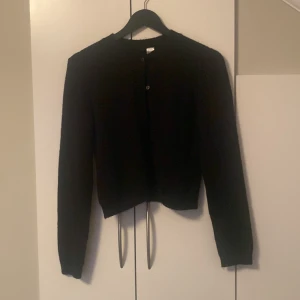 Svart kofta från Divided, storlek S - En svart, klassisk kofta från Divided i storlek S. Koftan har rund halsringning, knäppning framtill med små knappar och långa ärmar. Perfekt att slänga över en t-shirt eller linne för en enkel och stilren look.