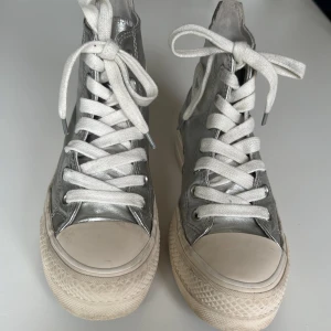 Converse Chuck Taylor All Star Canvas Silver - Snygga high top Converse sneakers i metallic silver med vita snören och vit sula. Skorna har klassisk rund tå och platt sula, med Converse-logga på sidan. Ovandelen är i syntetmaterial och insidan i textil. Perfekta för dig som vill sticka ut med en cool metallic-look. Nypris 1020 sek.