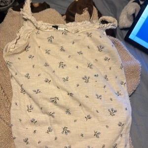 Blommig ribbad linnetopp från H&M Divided - Supersöt ljusbeige ribbad linnetopp från H&M Divided med små blå blommor och spetsdetaljer längs axelbanden och halsringningen. Perfekt för en chill sommardag eller när du vill ha en romantisk vibe. Tunn och mjuk bomullskänsla.