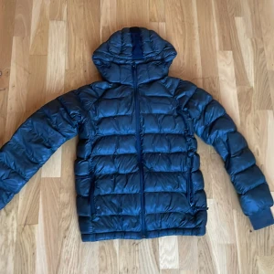 Mörk grön dunjacka Peak Performance - Snygg svart dunjacka från Peak Performance med huva och dragkedja framtill. Jackan har quiltad design, två sidofickor med dragkedja och diskret logga på bröstet. Perfekt för kalla dagar och riktigt skön att bära.