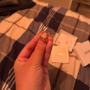 Guldig Dior ring med logga - Snygg ring från Dior i guldtonad metall med tydlig DIOR-logga framtill. Ringen har en stilren och modern design med små dekorativa detaljer längs bandet. Kommer med original ask och tygpåse med Dior-tryck. Perfekt statement piece för dig som gillar lyxiga accessoarer. Storlek M/52. Kommer nästan aldrig till användning. Nypris ca 3500kr, pris kan diskuteras