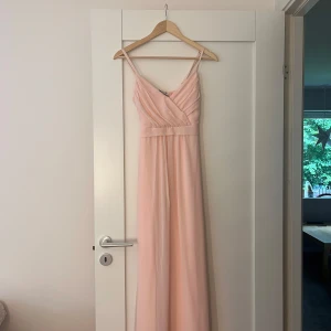 Ljusrosa långklänning med rosett - Superfin ljusrosa långklänning med smala axelband och omlottdetalj framtill. Klänningen har ett band i midjan som knyts till en söt rosett bak. Luftigt tyg som faller snyggt och ger en romantisk vibe. Perfekt för somriga tillfällen eller skolbal.