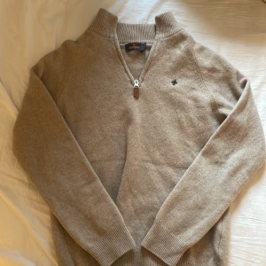Beige halvzip-tröja från Morris - Snygg beige stickad tröja från Morris med halv dragkedja och ribbad krage. Tröjan har lång ärm och en broderad logga på bröstet. Perfekt för lager-på-lager och passar till både jeans och chinos. Strl M sitter som XS