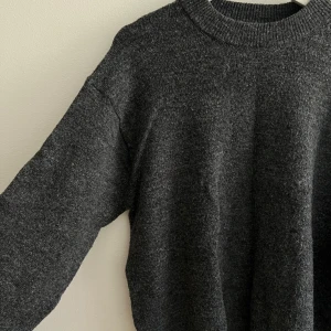 Mörkgrå stickad tröja med rund hals - Mysig mörkgrå stickad tröja med rund halsringning och långa ärmar. Perfekt för lager-på-lager och chill dagar. Tröjan har en enkel och stilren look med ribbade muddar vid hals, ärmslut och nederkant.