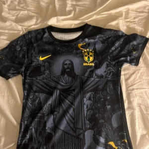 Svart Brasil Nike fotbollströja - Unik svart Brasilien fotbollströja från Nike med detaljerat tryck av Kristusstatyn och brasilianska motiv. Gula Nike-loggan och Brasil-märket på bröstet. Korta ärmar och rund hals. Materialet är lätt och andas, perfekt för match eller träning.