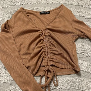Beige croppad topp med snörning Boohoo - Trendig croppad topp från Boohoo i beige ribbad bomullsmix. Långärmad med v-ringning och snygg snörning framtill som ger en rynkad effekt. Perfekt för dig som gillar en stilren men ändå cool look.