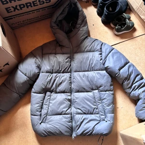 Grå pufferjacka från 157 med huva - Säljer en grå pufferjacka från 157 i storlek S. Jackan passar även M och L ifall man inte drar åt den. Jackan har en schysst huva, två snedställda fickor och dragkedja framtill. Perfekt för kalla dagar när du vill hålla värmen och ändå se clean ut.