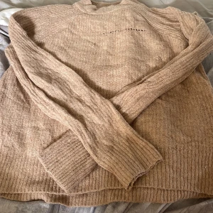 Beige stickad tröja med ribbade detaljer - Mysig beige stickad tröja från H&M Basic med ribbade muddar och krage. Tröjan har lång ärm och en relaxed fit, perfekt för lager på lager. Snygg strukturstickning och diskret hål i tröjan men om man har något under så syns det inte 