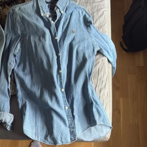 Ljusblå jeansskjorta Polo Ralph Lauren - Säljer en ljusblå jeansskjorta från Polo Ralph Lauren i slim fit, den är knappt använd. Skjortan har klassisk krage, knäppning framtill med vita knappar och lång ärm. Ikoniska broderade loggan på bröstet. Perfekt för en chill och stilren look.