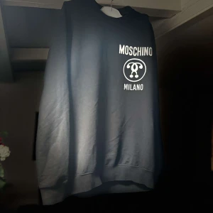 Svart sweatshirt från Moschino - Snygg svart sweatshirt från Moschino med stort vitt tryck på framsidan. Tröjan har långa ärmar och en klassisk rund halsringning. Perfekt för en stilren och avslappnad look.