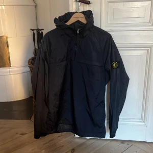 Mörkblå vindjacka från Stone Island - Säljer en snygg mörkblå vindjacka från Stone Island med huva och dragkedja. Jackan har en ikonisk logga på ärmen och är perfekt för blåsiga dagar. Den är både stilren och funktionell. Kom privat för att diskutera pris 🏆 😃